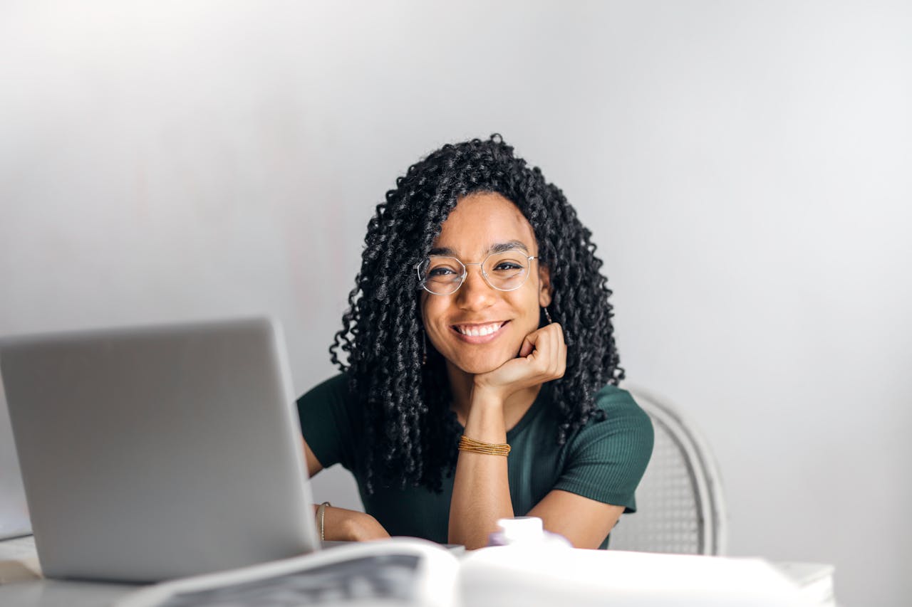 our-story Joyful businesswoman with curly hair smiling at camera while using laptop indoors.