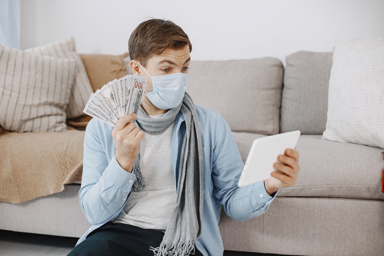 about-us A man in a face mask holding cash and using a tablet in a cozy living room setting.